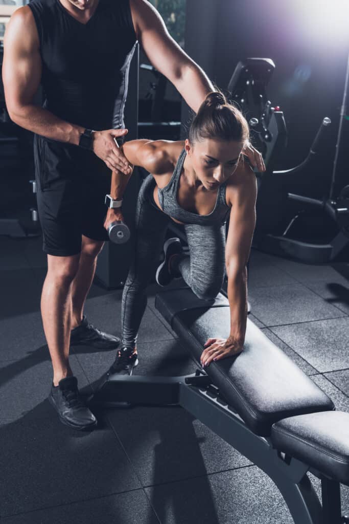 Une femme concentrée s'entraîne avec un haltère (4kg) sur un banc en salle de sport, aidée par son coach personnel.
