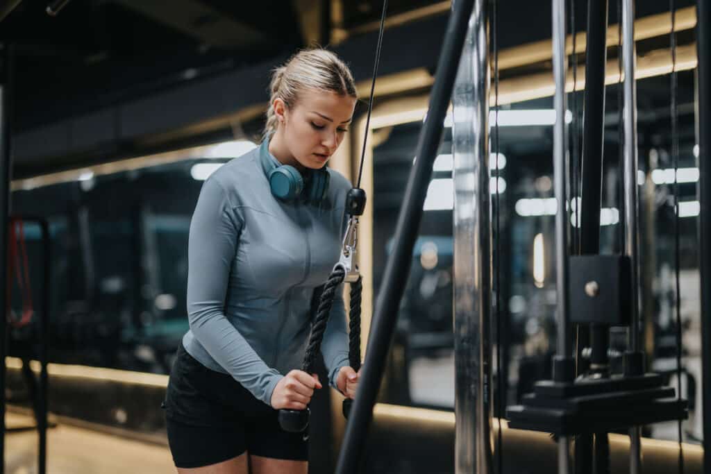 Jeune femme blonde concentrée s'entraîne sur machine à câble en salle de sport. Elle porte un haut bleu, short noir Nike et écouteurs bleus.