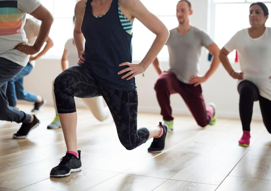 Groupe de personnes diversifiées en tenue de sport effectuant des fentes avant lors d'un cours de fitness en salle lumineuse.