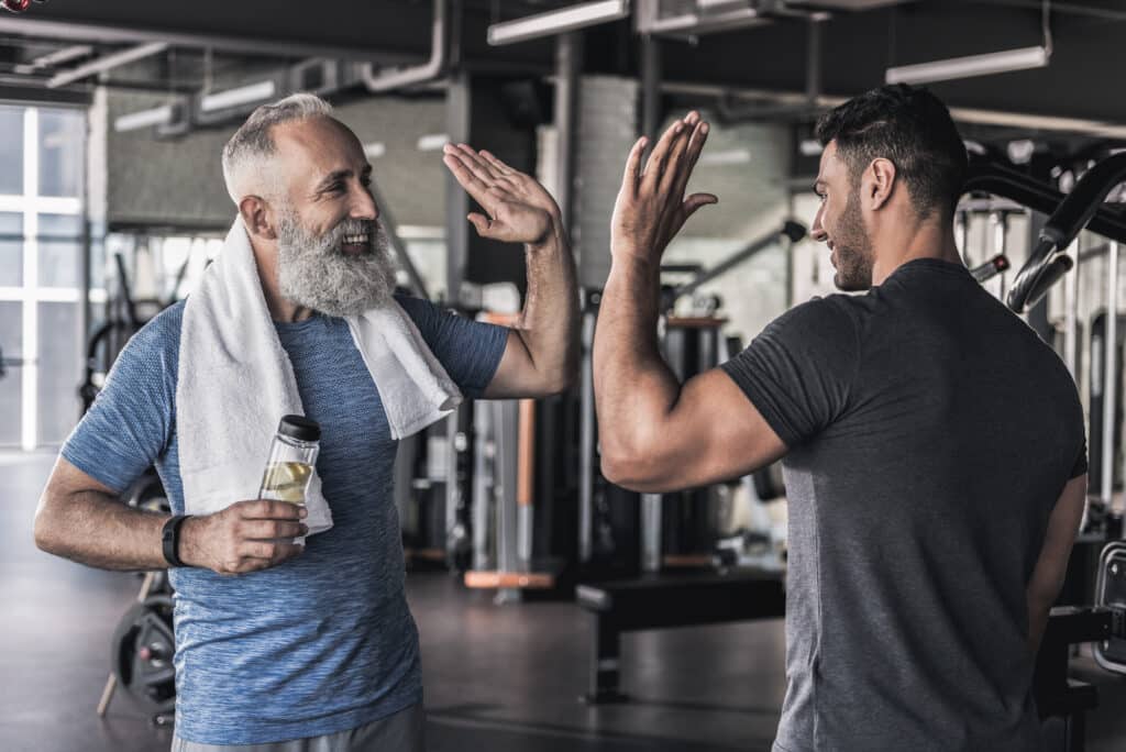 Fitness et amitié: Deux hommes se motivent à la salle. Deux hommes souriants se donnent un high-five dans une salle de sport. L'homme âgé barbu tient une bouteille d'eau.