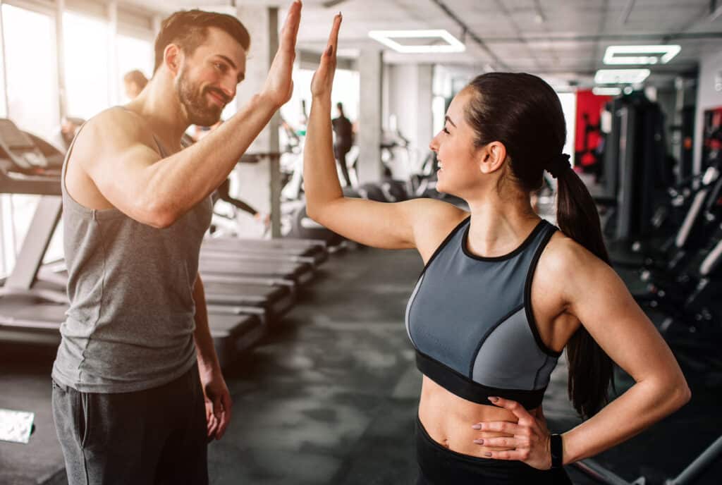 High-five motivant en salle de sport : amis célèbrent l'entraînement Deux amis souriants, un homme et une femme en tenue de sport, se donnent un high-five motivant dans une salle de sport moderne.