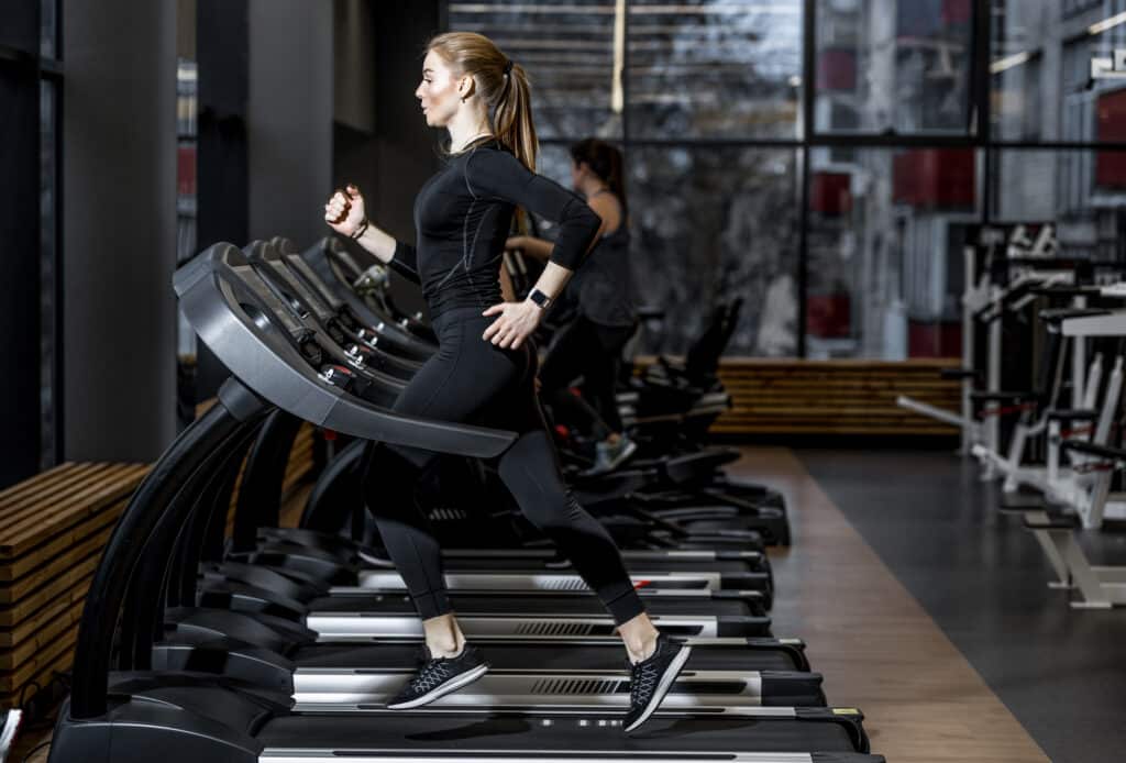 Entraînement Cardio : Course sur tapis de sport moderne Sportive en tenue noire, courant sur un tapis de course moderne. Salle de sport lumineuse avec fenêtres et équipements.
